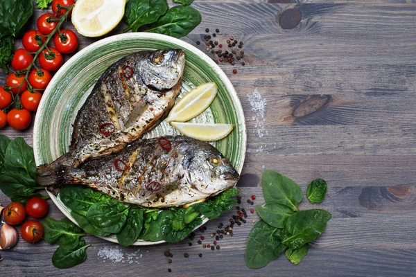 Dorado pescado horneado en el horno en las especias en un plato verde sobre un fondo de madera gris. Cerca de espinacas, limón, aceite de oliva, tomates cherry, pimienta — Foto de Stock