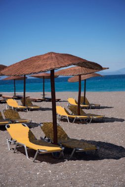 Rodos adasındaki plaj. Hasır ve sarı yatak, parlak mavi gökyüzü ve deniz şemsiyeleri. Seyahat ve tatil konsepti