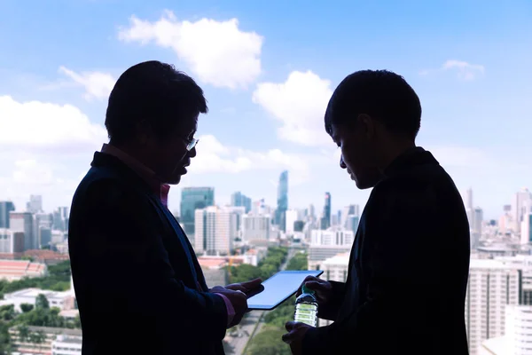 Silhouette of two executive businessmen discussing over a project on a computer tablet with city highrise buildings and sky view in the background, good for business team concept