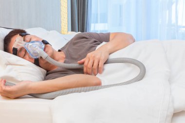 CPAP maske takıyor, hava hortumu için bağlanma, onun yatak odası ve geniş pencere, iyi uyku apnesi hikayesi için yatağında uyuyan Asyalı adam