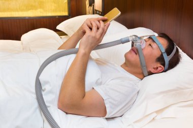 Orta yaş Asya giyen Cpap şapka adam ve hortum hava bağlanma ve onun yatak odasında yatakta döşeme sırasında telefonunu kullanarak maske