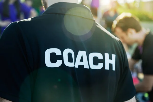 Sport coach in black shirt with white Coach text on the back standing outdoor at a school field, with morning lens flare, good for coaching or sport concept
