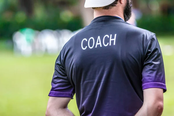 Back view of male soccer or football coach in dark shirt with word COACH written on back, standing on the sideline watching his team play, good for sport or coaching concept