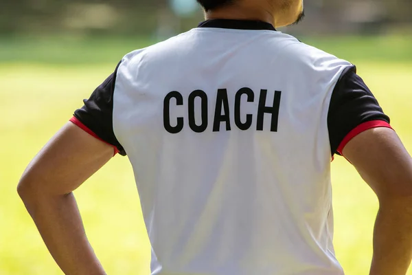 Back view of male soccer or football coach in white shirt with word COACH written on back, standing on the sideline watching his team play, good for sport or coaching concept