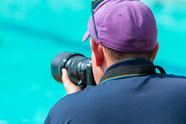 Back view of a male photographer taking picture with his camera at a swimming pool on a bright sunny day, blurred background of swimming pool water
