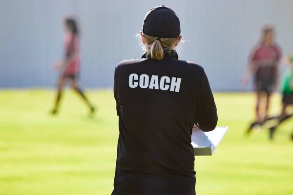 Back view of female sport coach in black COACH shirt at an outdoor sport field, watching her girl football team