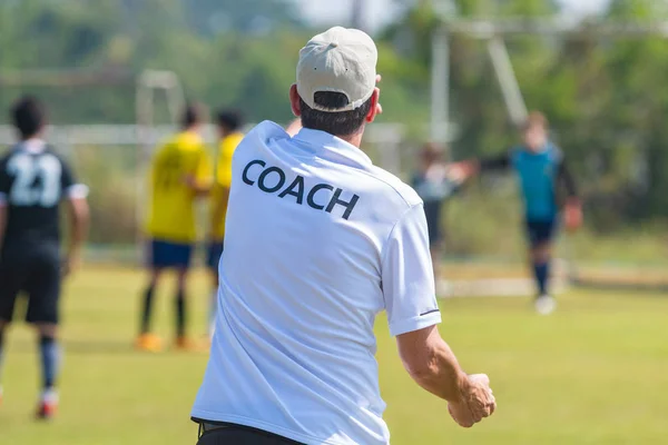 Back view of male football coach in white COACH shirt at an outdoor football field giving direction to his football team