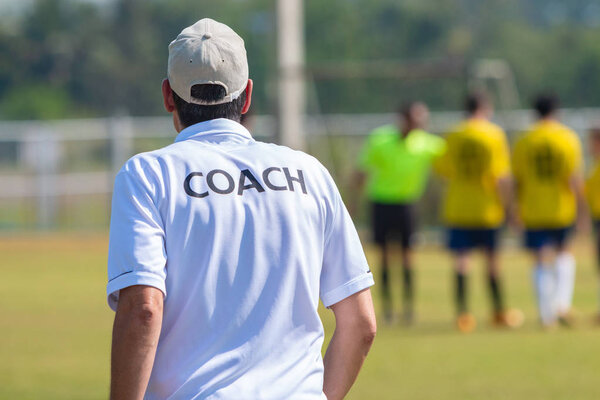 Back view of male football coach in white COACH shirt at an outdoor sport field, watching his team play on a sunny day
