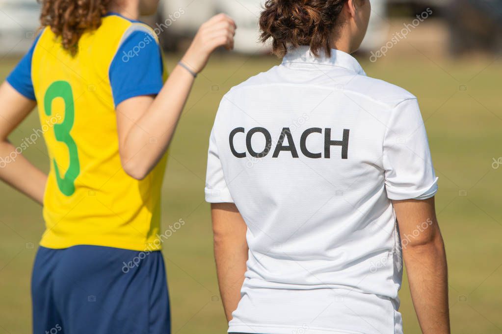 Back of sport coach wearing COACH shirt at an outdoor sport field, standing with a soccer player, watching the game
