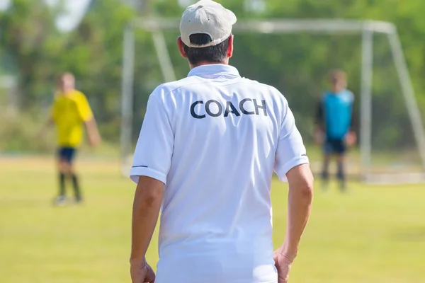 Back of football coach wearing white COACH shirt at an outdoor sport field coaching his team during a game, good for sport or coaching concept