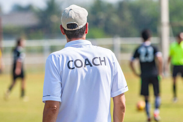 Back of male football coach wearing white COACH shirt at an outdoor sport field coaching his team during a game, good for sport or coaching concept