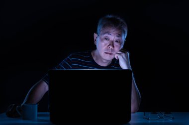 Yorgun orta yaş Asyalı erkek gece kendi ev ofisinde kendi bilgisayar üzerinde çalışıyor