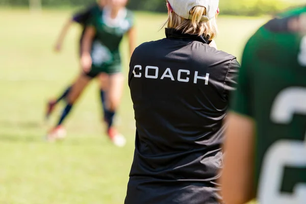 Vista trasera de una mujer de fútbol, fútbol, entrenador en camisa de entrenador negro de pie en el sol viendo a su equipo jugar en un campo de fútbol al aire libre — Foto de Stock