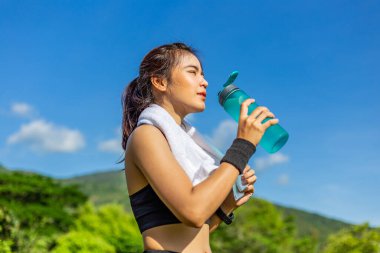 Güzel, genç Asyalı bir kadın sabah koşu pistinde egzersiz yapıyor. Su içmek için dinleniyor. Parlak güneşli bir günde, masmavi bir arka plan.