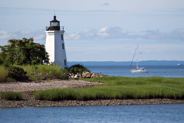 Sailboat passing by Black Rock Harbor lighthouse, also referred to as Fayerweather Island light, near the rocky shoreline on a warm summer day.