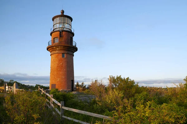 Tahta çitler Massachusetts 'teki Marthas Vineyard Adası' nda bir yaz günü Gay Head feneri olarak da bilinen Aquinnah deniz fenerinin büyük tuğla kulesine çıkar. Batan güneş kuleyi aydınlatıyor..
