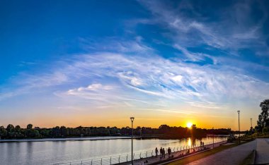 Nehir kenarında romantik bir günbatımı. Tüylü bulutların sakin mavi gökyüzündeki manzarası. Sarı gün batımı, yaya geçidinde geçenleri rahatlatıyor. Güzel, yumuşak bir akşam yürüyüşü..