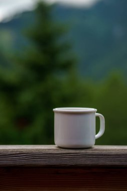 Orman ve dağların arka planında içkili beyaz bir kupa görüntüsü. Sıcak açık hava rahatlığı