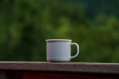 Orman ve dağların arka planında içki içen küçük beyaz bir kupa. Sıcak açık hava rahatlığı