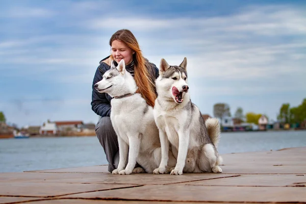 Молодая красивая девушка с рыжими волосами сидит на пирсе с двумя хаски-собаками. Женщина с парой сибирских хаски — стоковое фото