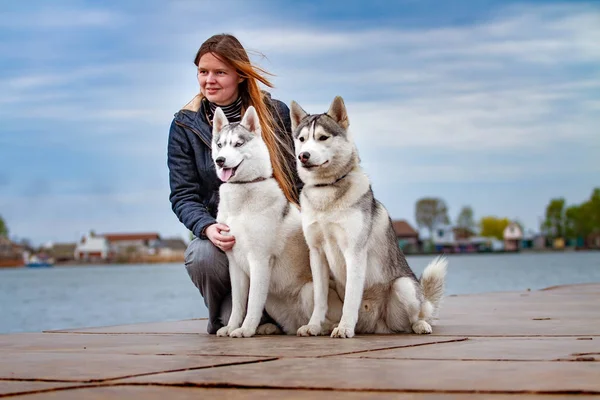 Young beautiful girl with red hair is sitting on a pier with two husky dogs. Woman with a pair of Siberian Husky — Stock Photo, Image