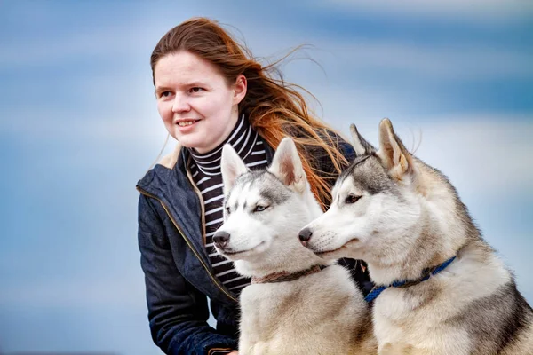 Joven chica hermosa con el pelo rojo está sentado en un muelle con dos perros husky. Mujer con un par de Husky siberiano —  Fotos de Stock