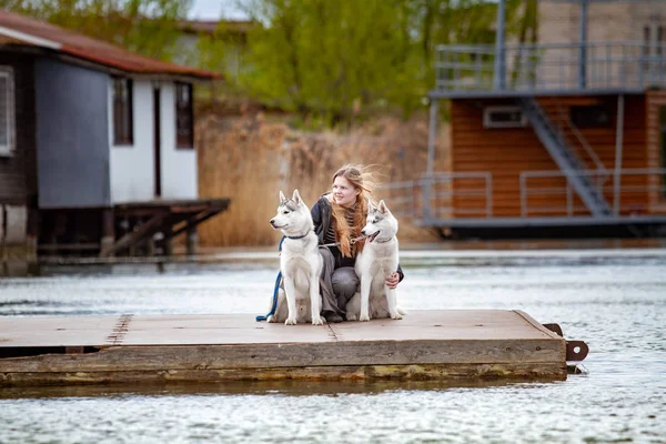 Young beautiful girl with red hair is sitting on a pier with two husky dogs. Woman with a pair of Siberian Husky