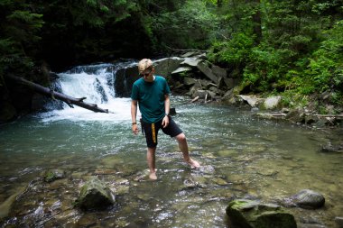 Genç adam nehrin kenarında bir şelalenin arka planında duruyor.