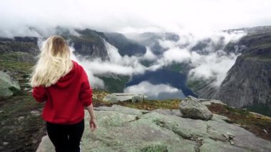 Kırmızı bir kazak uzun saçlı genç bir kız dağlarda yürür. Norveç Fiyordları.