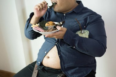 şişman adam kilolu kişinin portre yemek, aç hissediyor ve izole kırpma yolu ile beyaz arka plan üzerinde yemek fişleri, kek, armchai üzerinde oturmuş yeşil çay frappe