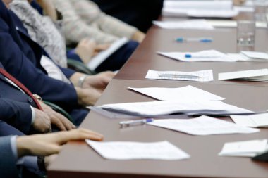 Çerçevede, insanlar bir masada oturuyor, yüzleri görünmüyor. İş kağıtları ve kalemler masada.