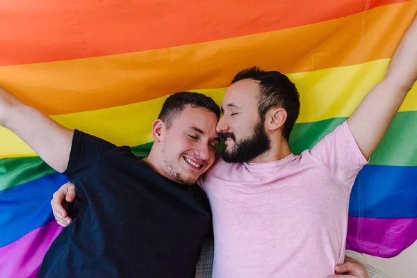 Birbirini tutan ve LGBTQ bayrağı taşıyan iki beyaz homoseksüel adamın stok fotoğrafı. Gözleri kapalı..