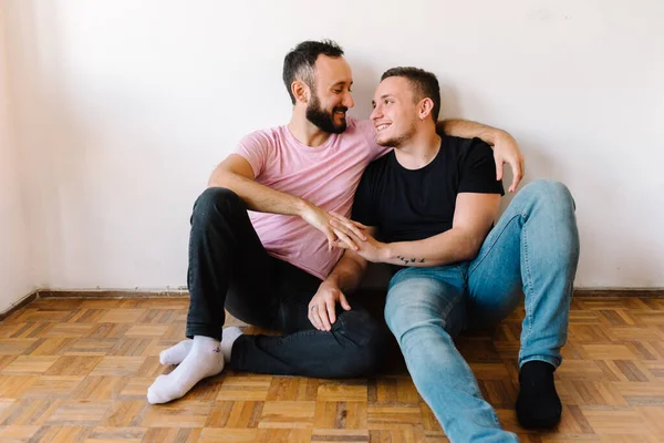 Stock photo of two caucasian homosexual men sitting on the floor in their apartment. One of them is hugging the other one from behind.