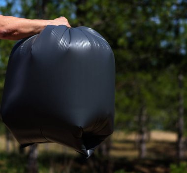 elini karşı bir orman çöp dolu tutan büyük bir siyah plastik torba, bir yaz günü, çöp toplama doğada