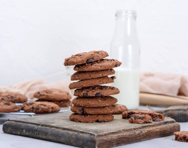 Kahverengi tahta üzerinde yuvarlak çikolatalı kurabiye yığını, bir şişe sütün arkasında.