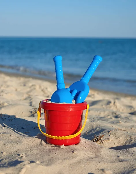 Детское красное пластиковое ведро с песком и лопатой на берегу — стоковое фото