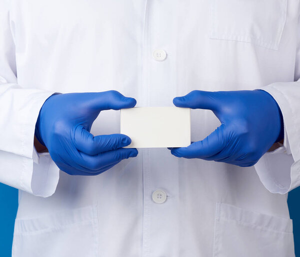 doctor dressed in a white robe uniform and blue sterile gloves is standing and holding empty white paper business card, place for inscription