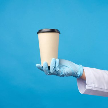 Mavi lateks bir eldiven içinde kağıt bir kullan-at karton bardak ve plastik kapaklı mavi arka planda, salgın sırasında güvenli ve temas kurulamayan siparişler sunar.