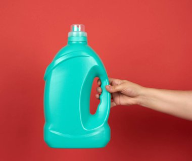 El, kırmızı arka planda çamaşır yıkamak için sıvı deterjan içeren yeşil büyük bir şişe tutuyor.