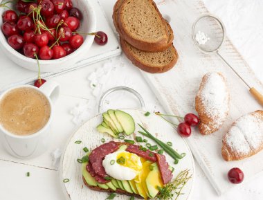 Yuvarlak bir tahtanın üzerinde yumurta ve avokado ile kızarmış ekmek, kruvasan ve olgun kırmızı kirazların yanında, sabah kahvaltısı, beyaz bir masanın üstünde üst manzara.