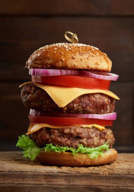 Domatesli duble çizburger, soğan, barbekü pirzolası ve eski bir tahta kesme tahtasında susamlı ekmek, kahverengi arka plan. Fast food