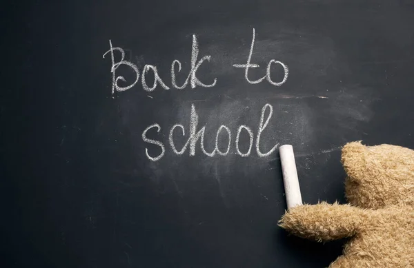 lettering back to school in white chalk on a black school board and a piece of chalk in the paw of a brown teddy bear