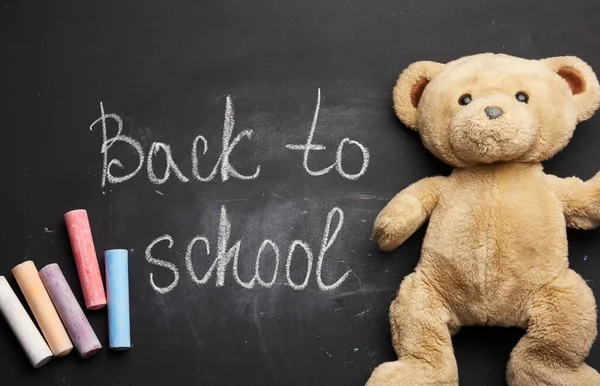 lettering back to school in white chalk on a black school board and a piece of chalk, brown teddy bear
