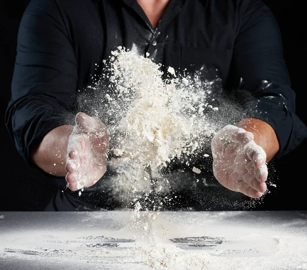 黒の制服のシェフが別の方向に白い小麦粉を振りかける 製品散乱ほこり 黒の背景 テーブルに座っている男 — ストック写真