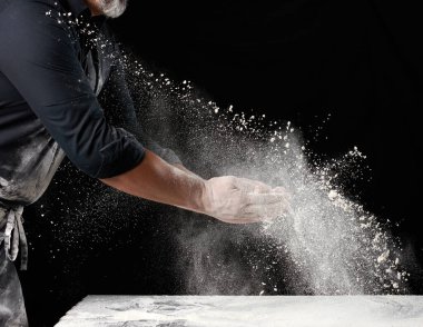 Siyah üniformalı şef beyaz buğday ununu farklı yönlere serper, ürün tozları dağıtır, siyah zemin, parçacıklar dağılır ve havada donar.