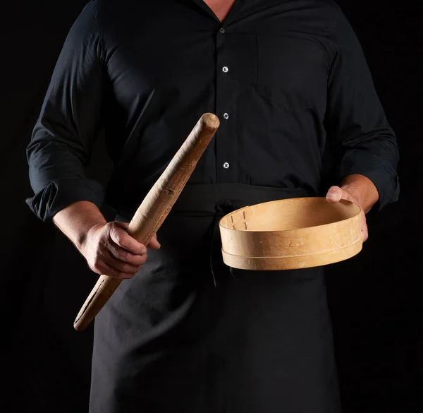 man in black uniform holding empty vintage round wooden sieve for sifting flour and rolling pin, chef stands against black background