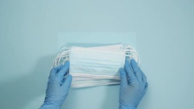Mavi lateks eldivenli iki el mavi arka planda yatıyordu. Tek kullanımlık tıbbi maske yığını, virüslere ve bakterilere karşı koruma, üst görünüm.