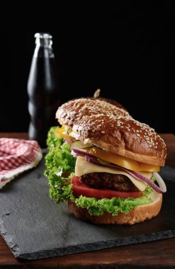 Lezzetli hamburgerler, kızarmış sığır pirzolası, soğan, domates ve susamlı beyaz buğday unlu çörek. Ahşap bir tahtanın üzerinde, bir şişenin arkasında, bir içecekle.