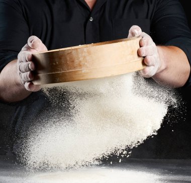 Şef siyah üniformalı bir adam elinde yuvarlak bir ahşap elek tutar ve siyah arka planda beyaz buğday unu seçer, parçacıklar farklı yönlere uçar, tozlu uzay