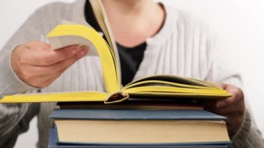 Gri elbiseli kadın büyük bir kitap yığınının üzerinde oturuyor, elinde sarı bir kitap, sayfaları çeviren bir kadın, beyaz arka plan, yavaş çekim